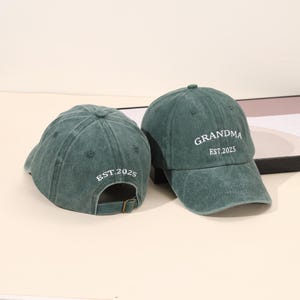 May include: Two green baseball caps. One cap has the word "GRANDMA" and "EST.2025" printed in white. The other cap has "EST.2025" printed in white. Both caps have an adjustable strap.