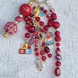 May include: A gold-toned keychain featuring a variety of red beads in different shapes and textures, including faceted, smooth, and glittery finishes. Charms include a colorful ABC block, a stack of books, a pencil, and an apple with the word Teacher.