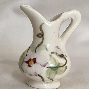 May include: A small, cream-colored ceramic pitcher featuring a hand-painted floral design. The pitcher has a rounded body, a flared spout, and a curved handle. The floral pattern consists of white petals with purple accents, a yellow center, and green vines.