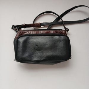 Pode incluir: Bolsa transversal de couro preto com uma se&ccedil;&atilde;o superior marrom texturizada. A bolsa tem uma al&ccedil;a longa e ajust&aacute;vel e uma al&ccedil;a mais curta. A bolsa &eacute; retangular e possui fecho de z&iacute;per.