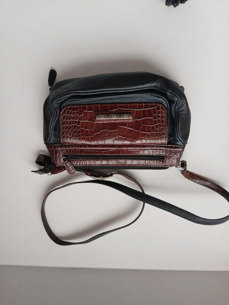 Pode incluir: Bolsa transversal preta e marrom com um bolso frontal de pele de crocodilo sint&eacute;tica. A bolsa tem uma parte superior de couro preto e uma al&ccedil;a longa e ajust&aacute;vel. A bolsa tem fecho de z&iacute;per e um pequeno la&ccedil;o na lateral.