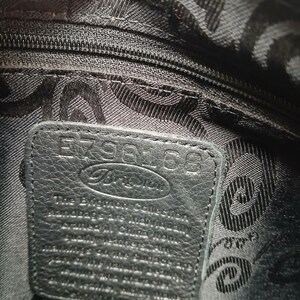 Pode incluir: Vista de perto do interior de uma mala de m&atilde;o preta. Uma etiqueta de couro com a marca "Brighton" e o n&uacute;mero "E798168" &eacute; vis&iacute;vel. O forro tem um padr&atilde;o repetido.