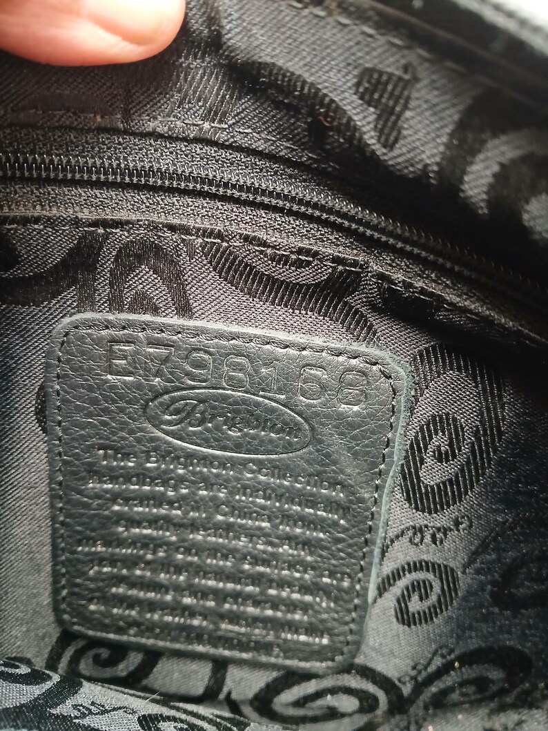 Pode incluir: Vista de perto do interior de uma mala de m&atilde;o preta. Uma etiqueta de couro com a marca "Brighton" e o n&uacute;mero "E798168" &eacute; vis&iacute;vel. O forro tem um padr&atilde;o repetido.
