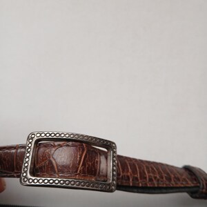Pode incluir: Close-up de um cinto de couro castanho com uma fivela prateada. O cinto tem um padr&atilde;o texturizado, semelhante ao de um crocodilo. A fivela retangular tem uma borda decorativa.