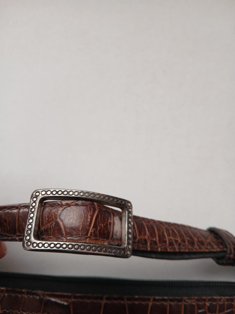 Pode incluir: Close-up de um cinto de couro castanho com uma fivela prateada. O cinto tem um padr&atilde;o texturizado, semelhante ao de um crocodilo. A fivela retangular tem uma borda decorativa.