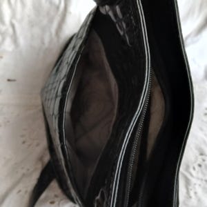 以下が含まれることがあります： 光沢のある質感の黒いハンドバッグ。ファスナー付きのコンパートメントと広々とした内部を備えています。白いトリムが縁に沿って施され、コントラストのあるディテールを加えています。バッグは白い質感の背景に置かれています。