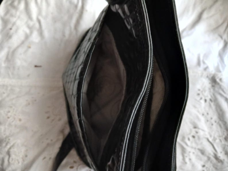 以下が含まれることがあります： 光沢のある質感の黒いハンドバッグ。ファスナー付きのコンパートメントと広々とした内部を備えています。白いトリムが縁に沿って施され、コントラストのあるディテールを加えています。バッグは白い質感の背景に置かれています。