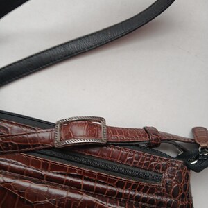 Pode incluir: Bolsa transversal de couro castanho com padr&atilde;o de crocodilo e al&ccedil;a de couro preta. A bolsa apresenta uma fivela e z&iacute;per prateados. A bolsa tem uma superf&iacute;cie texturizada e um detalhe em couro preto.