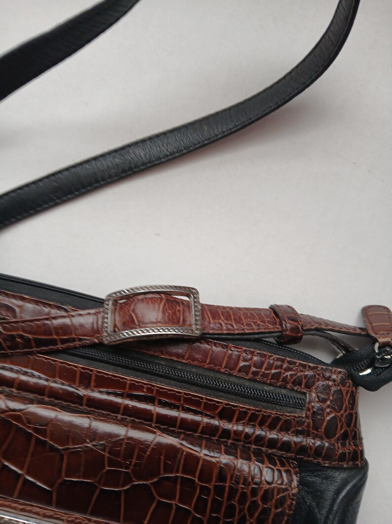 Pode incluir: Bolsa transversal de couro castanho com padr&atilde;o de crocodilo e al&ccedil;a de couro preta. A bolsa apresenta uma fivela e z&iacute;per prateados. A bolsa tem uma superf&iacute;cie texturizada e um detalhe em couro preto.