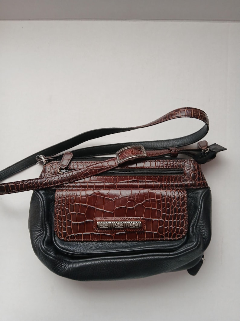 Pode incluir: Bolsa transversal preta e castanha com um design texturizado de imita&ccedil;&atilde;o de pele de crocodilo. A bolsa apresenta um bolso frontal castanho e uma al&ccedil;a longa e ajust&aacute;vel. Uma placa de metal prateada com texto est&aacute; na frente.