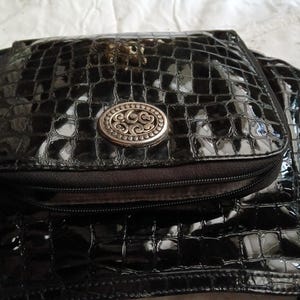以下が含まれることがあります： シルバーの楕円形のエンブレムが付いた、黒色の光沢のあるフェイククロコダイルスキンハンドバッグ。バッグにはジッパー付きポケットとフラップクロージャーが付いています。バッグにはショルダーストラップとタッセルのディテールがあります。バッグはスタイリッシュなアクセサリーです。