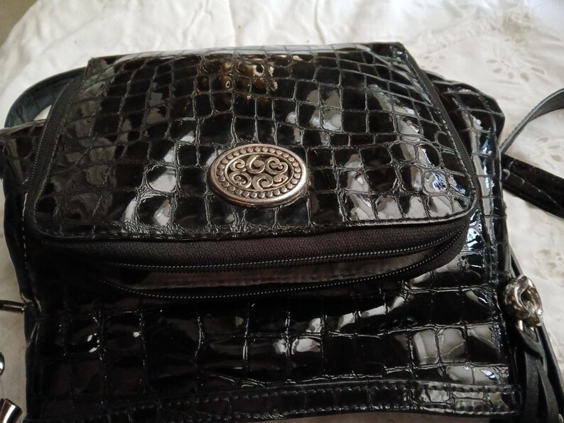 以下が含まれることがあります： シルバーの楕円形のエンブレムが付いた、黒色の光沢のあるフェイククロコダイルスキンハンドバッグ。バッグにはジッパー付きポケットとフラップクロージャーが付いています。バッグにはショルダーストラップとタッセルのディテールがあります。バッグはスタイリッシュなアクセサリーです。