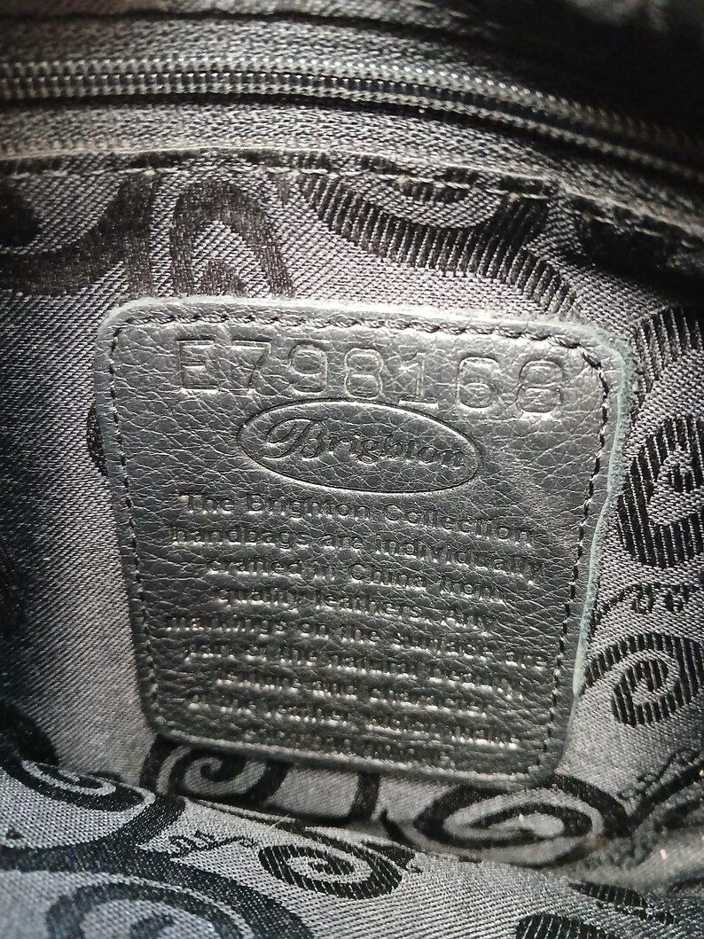 Pode incluir: Close-up do interior de uma mala de m&atilde;o de couro preto. Uma etiqueta retangular de couro &eacute; costurada no interior, com a marca "Brighton" e o n&uacute;mero de s&eacute;rie E798168 em relevo. O forro apresenta um padr&atilde;o repetitivo.