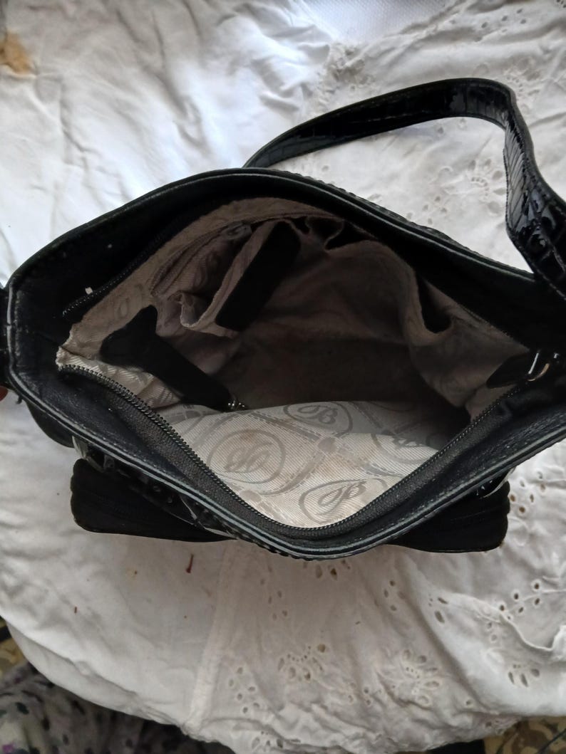 以下が含まれることがあります： 黒い革のハンドバッグ。表面はテクスチャ加工、持ち手は光沢があります。内側には複数のコンパートメントと模様入りの裏地があります。ジッパー開閉で、必需品を持ち運ぶように設計されています。