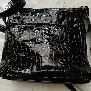 以下が含まれることがあります： クロコダイル柄の黒い光沢のあるクロスボディバッグ。バッグには、調節可能な長いストラップとシルバーカラーのバックルが付いています。バッグは長方形で、背面に小さなポケットが付いています。