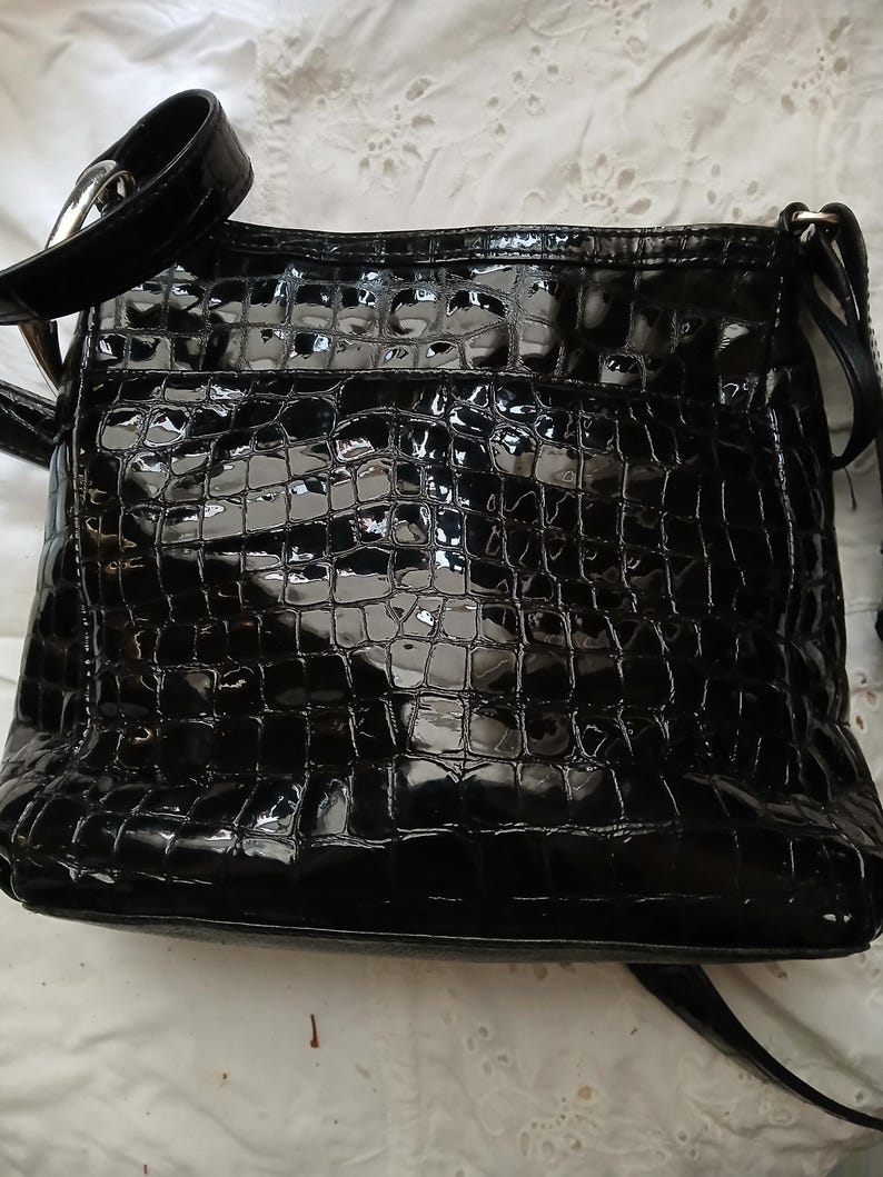 以下が含まれることがあります： クロコダイル柄の黒い光沢のあるクロスボディバッグ。バッグには、調節可能な長いストラップとシルバーカラーのバックルが付いています。バッグは長方形で、背面に小さなポケットが付いています。
