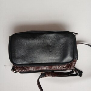 Pode incluir: Bolsa transversal de couro preto com base marrom texturizada com efeito de crocodilo. A bolsa tem uma al&ccedil;a longa e ajust&aacute;vel e um z&iacute;per prateado. A bolsa tem formato retangular.