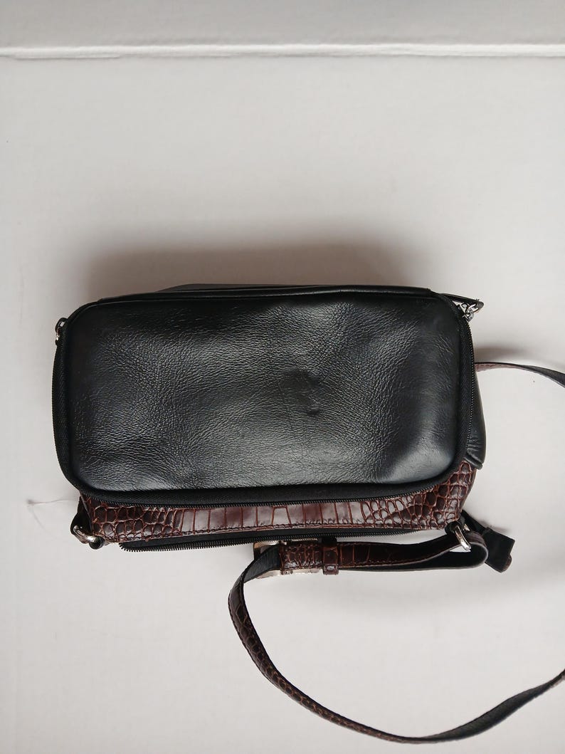 Pode incluir: Bolsa transversal de couro preto com base marrom texturizada com efeito de crocodilo. A bolsa tem uma al&ccedil;a longa e ajust&aacute;vel e um z&iacute;per prateado. A bolsa tem formato retangular.