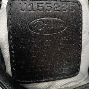 以下が含まれることがあります： ブランド名「Brighton」と番号「U155285」がエンボス加工された黒革のタグのクローズアップ。タグは、バッグの内側と思われる、質感のある明るい色の生地に縫い付けられています。