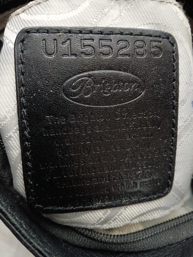 以下が含まれることがあります： ブランド名「Brighton」と番号「U155285」がエンボス加工された黒革のタグのクローズアップ。タグは、バッグの内側と思われる、質感のある明るい色の生地に縫い付けられています。