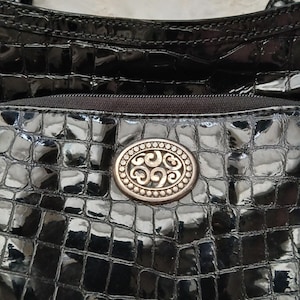 以下が含まれることがあります： 光沢のある質感の黒いハンドバッグ。バッグには、装飾的なデザインの楕円形の金属製エンブレムが付いています。バッグの上部にはジッパーが見えます。バッグの表面はワニのような模様です。