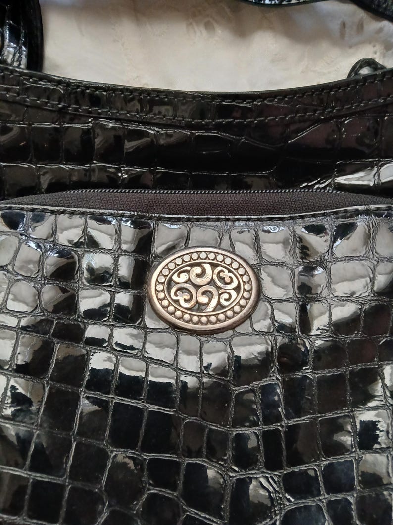 以下が含まれることがあります： 光沢のある質感の黒いハンドバッグ。バッグには、装飾的なデザインの楕円形の金属製エンブレムが付いています。バッグの上部にはジッパーが見えます。バッグの表面はワニのような模様です。