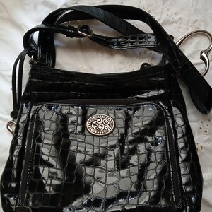 以下が含まれることがあります： 光沢のある黒いハンドバッグ。クロコダイルのような質感で、シルバーカラーの装飾的な留め金と、調節可能な長いストラップが付いています。前面にはファスナー付きポケットがあり、ハート型のハンドルアタッチメントが付いています。