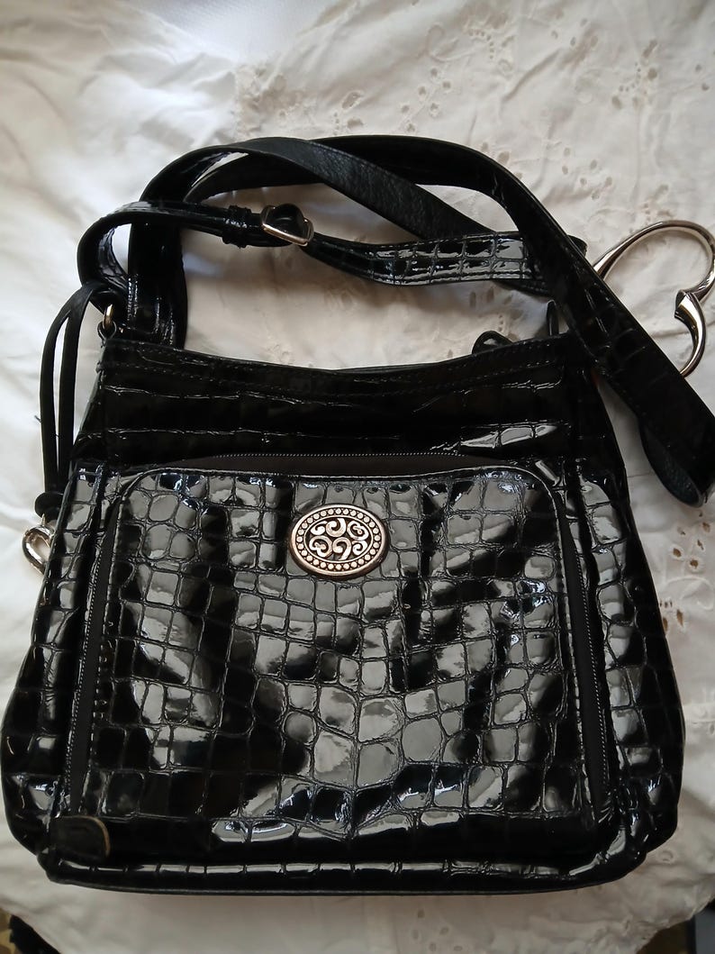 以下が含まれることがあります： 光沢のある黒いハンドバッグ。クロコダイルのような質感で、シルバーカラーの装飾的な留め金と、調節可能な長いストラップが付いています。前面にはファスナー付きポケットがあり、ハート型のハンドルアタッチメントが付いています。