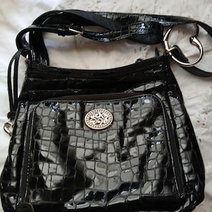 以下が含まれることがあります： 光沢のあるクロコダイル風の質感の黒いクロスボディバッグ。バッグには、装飾的なシルバーのエンブレムが付いたフロントポケットと、調節可能なストラップが付いています。バッグのストラップには、ハート型のシルバーリングが付いています。