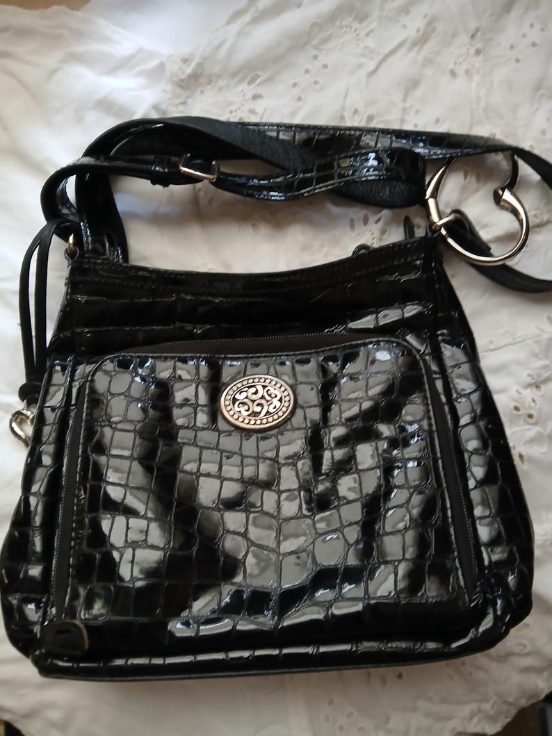 以下が含まれることがあります： 光沢のあるクロコダイル風の質感の黒いクロスボディバッグ。バッグには、装飾的なシルバーのエンブレムが付いたフロントポケットと、調節可能なストラップが付いています。バッグのストラップには、ハート型のシルバーリングが付いています。
