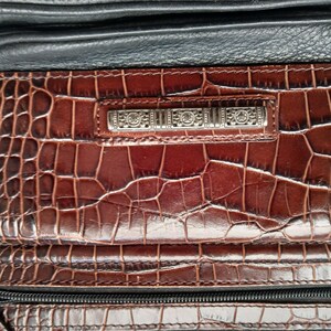 Pode incluir: Close-up de uma mala de m&atilde;o de couro castanho e preto. A mala apresenta um padr&atilde;o em relevo semelhante ao de um crocodilo na frente e uma barra decorativa prateada. Um z&iacute;per e uma al&ccedil;a pretos tamb&eacute;m s&atilde;o vis&iacute;veis. A mala parece ser uma mala de ombro ou tiracolo.