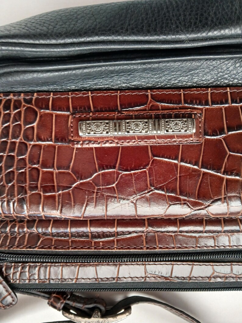 Pode incluir: Close-up de uma mala de m&atilde;o de couro castanho e preto. A mala apresenta um padr&atilde;o em relevo semelhante ao de um crocodilo na frente e uma barra decorativa prateada. Um z&iacute;per e uma al&ccedil;a pretos tamb&eacute;m s&atilde;o vis&iacute;veis. A mala parece ser uma mala de ombro ou tiracolo.