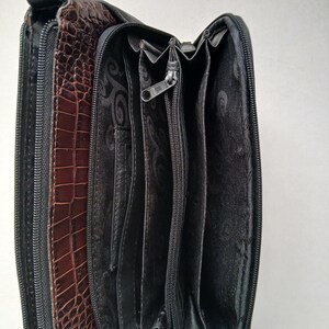 Pode incluir: Uma carteira de couro marrom e preta com textura de pele de crocodilo. A carteira est&aacute; aberta, revelando v&aacute;rios compartimentos e um fecho de z&iacute;per. O interior &eacute; preto com um design estampado.