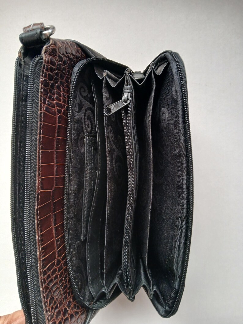 Pode incluir: Uma carteira de couro marrom e preta com textura de pele de crocodilo. A carteira est&aacute; aberta, revelando v&aacute;rios compartimentos e um fecho de z&iacute;per. O interior &eacute; preto com um design estampado.