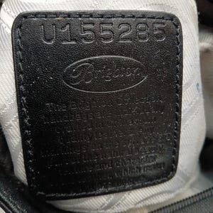以下が含まれることがあります： ブランド名「Brighton」と番号「U155285」がエンボス加工された黒い革製のタグのクローズアップ。タグは白い布の裏地の上に縫い付けられており、バッグのジッパーが見えます。