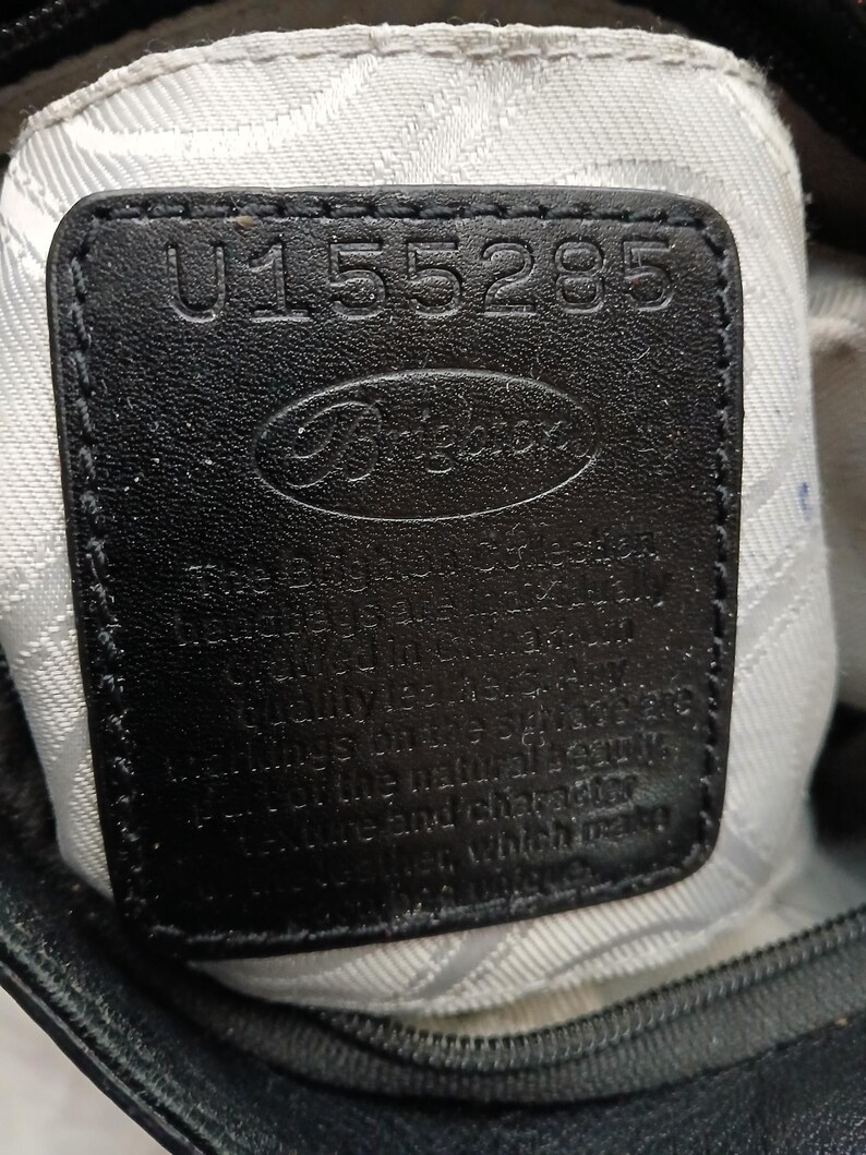 以下が含まれることがあります： ブランド名「Brighton」と番号「U155285」がエンボス加工された黒い革製のタグのクローズアップ。タグは白い布の裏地の上に縫い付けられており、バッグのジッパーが見えます。