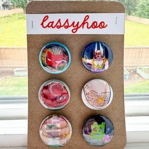 May include: Six round button pins with various cartoon characters and designs. The pins are displayed on a brown cardboard backing with the word "lassyhoo" printed in red.