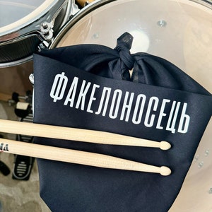 May include: Black fabric drum mufflers with the text "Факелоносець" in white lettering. Two wooden drumsticks are lying on top of the mufflers.