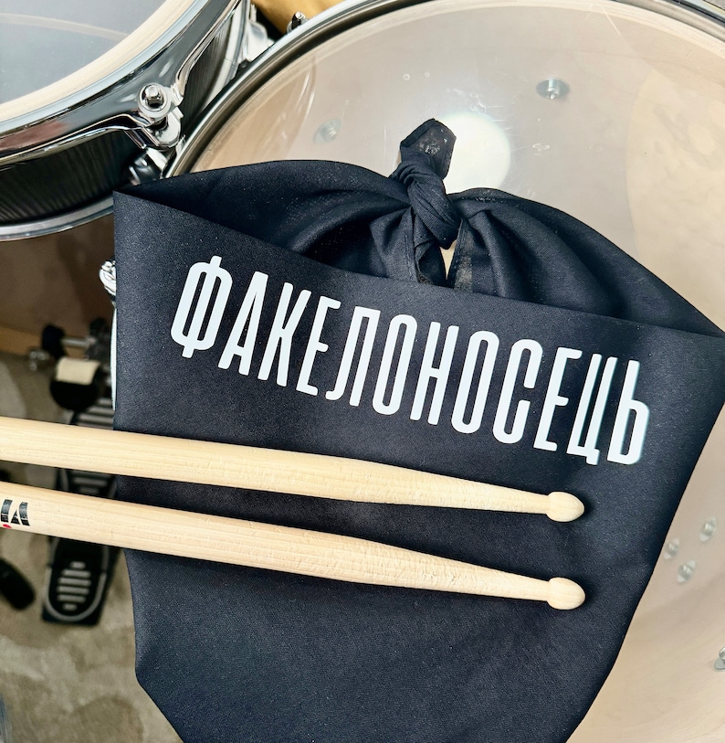 May include: Black fabric drum mufflers with the text "Факелоносець" in white lettering. Two wooden drumsticks are lying on top of the mufflers.