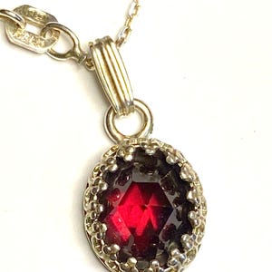 May include: A silver chain necklace with a red gemstone pendant. The pendant is oval shaped and has a silver crown-like setting.
