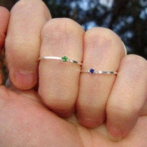 May include: Three silver rings with small gemstones. The ring on the left has a green gemstone, the middle ring has a blue gemstone, and the ring on the right has a clear gemstone.