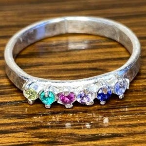 May include: A silver ring with six colorful gemstones. The gemstones are arranged in a row and include yellow, teal, magenta, purple, blue, and indigo. The ring is set on a wooden surface.
