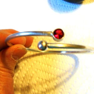 Puede incluir: Un brazalete de plata con una piedra roja y una perla de plata. El brazalete está sobre una superficie blanca.