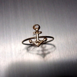 Puede incluir: Un anillo dorado con un diseño de ancla. El ancla es un diseño simple y estilizado con una parte superior redondeada y una base ligeramente curvada.