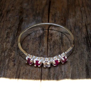 May include: A silver ring with a row of five red gemstones and a single clear gemstone in the center.