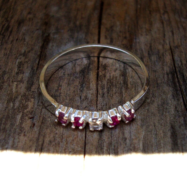 May include: A silver ring with a row of five red gemstones and a single clear gemstone in the center.