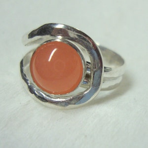 Può includere: Un anello d'argento con una grande pietra preziosa rotonda di colore pesca incastonata in un design a cerchio aperto testurizzato.