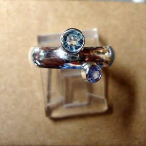 May include: A silver ring featuring two gemstones. The larger gemstone is a light blue aquamarine, and the smaller gemstone is a light purple amethyst.