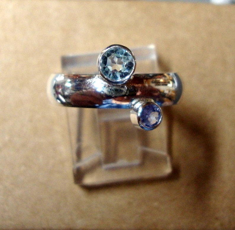 May include: A silver ring featuring two gemstones. The larger gemstone is a light blue aquamarine, and the smaller gemstone is a light purple amethyst.