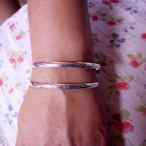 May include: Two silver bangles on a person's wrist. The bangles are simple and have a smooth, polished finish.